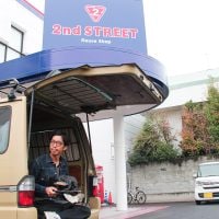 店舗をバックに自身の愛車と共に購入品をもつヘンミさん